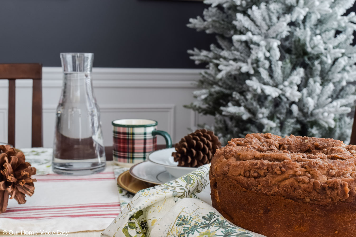 A round coffee cake sits on a festive table next to pine cones, plates, a plaid mug, a glass water carafe, and a snow-frosted artificial tree in the background.