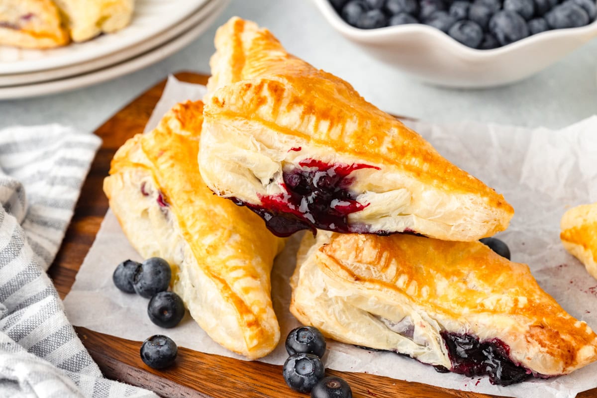Three golden-brown blueberry turnovers are stacked on parchment paper, surrounded by fresh blueberries, with a bowl of blueberries and plates visible in the background.