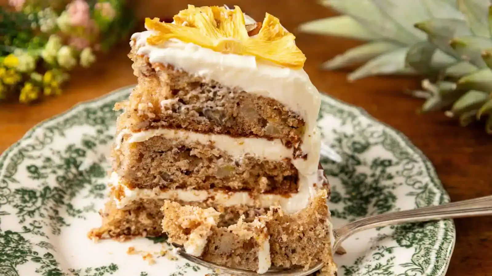 A slice of layered cake with cream frosting and pineapple garnish on a decorative plate, with a fork and blurred flowers in the background.