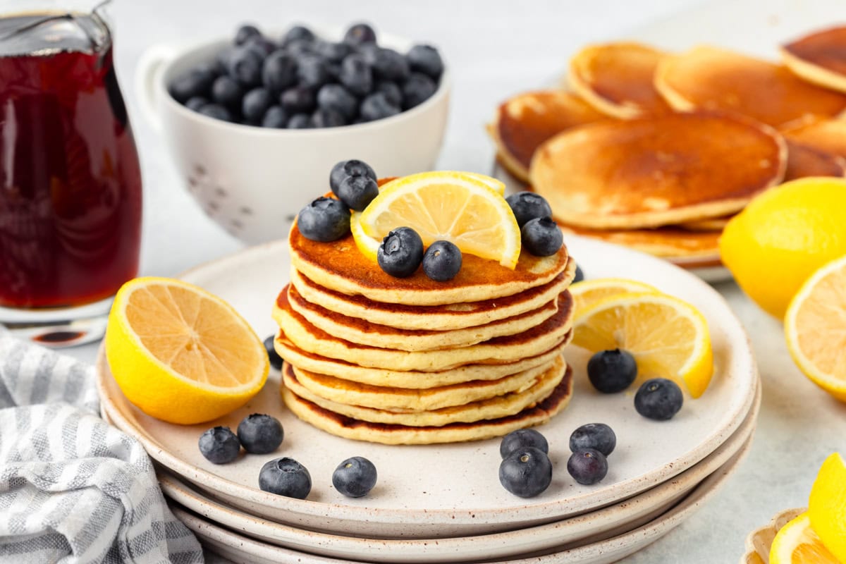 A stack of pancakes garnished with lemon slices and blueberries, surrounded by more pancakes, fresh blueberries, lemons, and a glass jug of syrup on a table.
