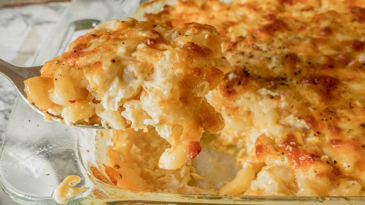 A close-up of a serving of baked macaroni and cheese being lifted from a glass baking dish, showing melted cheese and browned top.