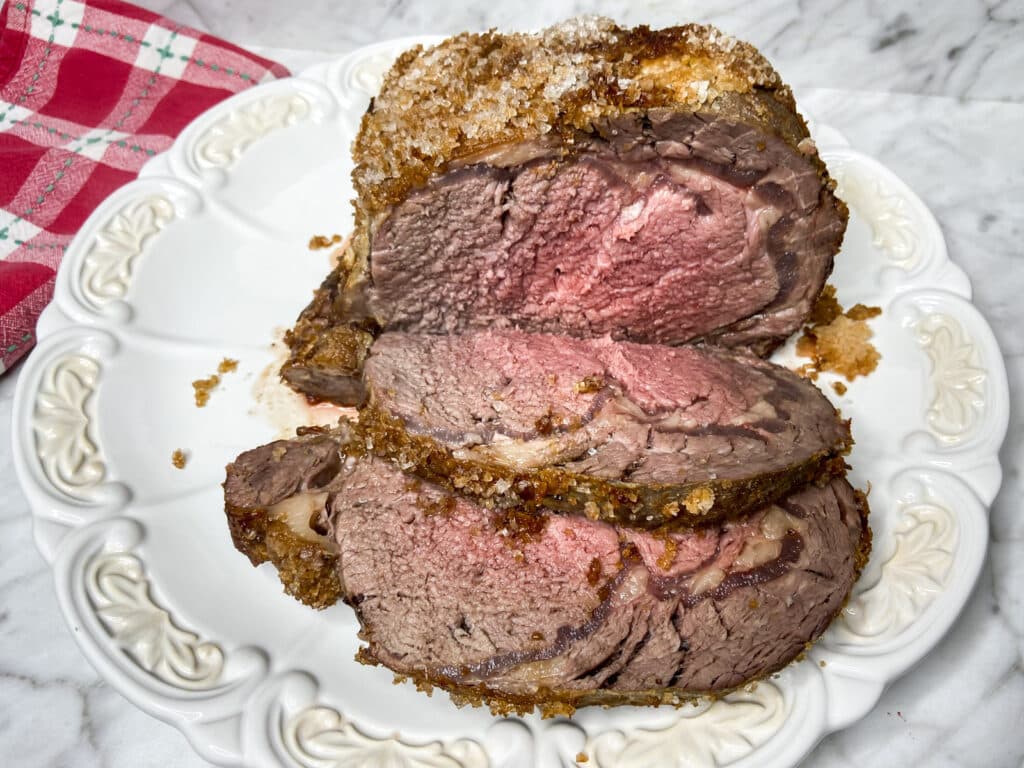 Sliced roast beef with a crispy crust is served on a white plate, with a red and white checkered cloth nearby.