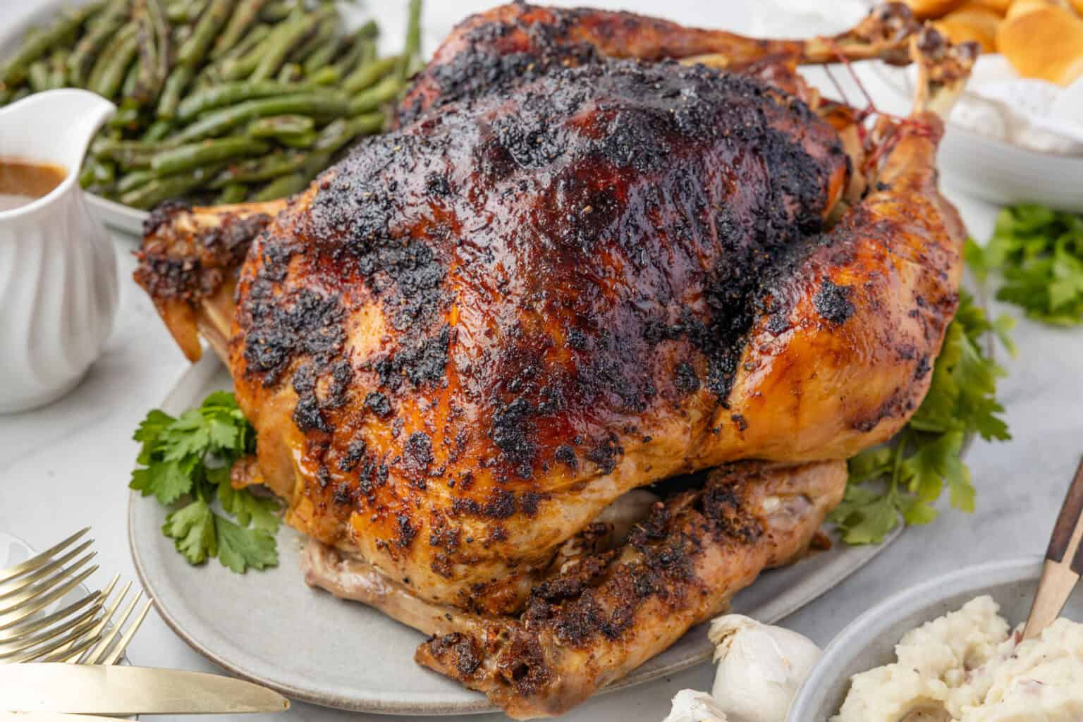 A whole roasted turkey with a dark, seasoned crust on a platter, surrounded by green beans, gravy, and other side dishes.