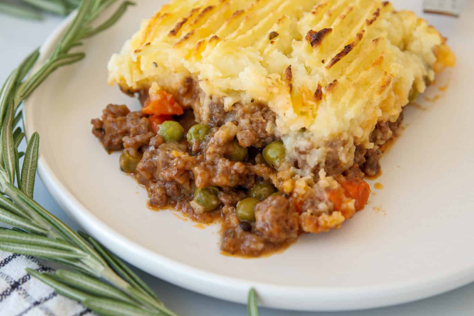 A serving of shepherdโs pie on a white plate, showing a layer of mashed potatoes on top of ground meat, peas, and carrots.