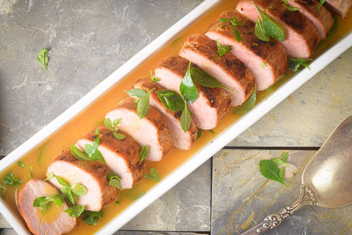Sliced pork tenderloin garnished with fresh herbs, served in a rectangular white dish with sauce, next to a metal serving spoon.