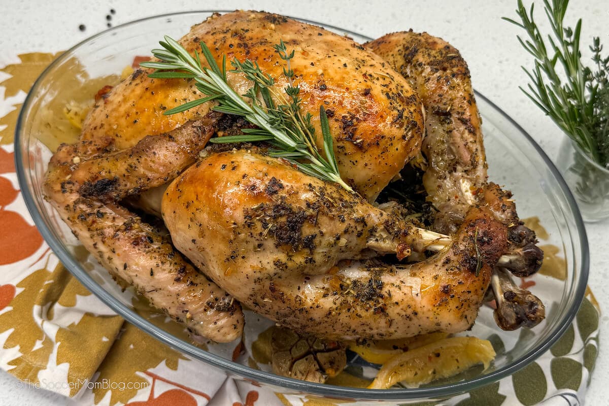 Roast chicken with herbs and lemon wedges in a glass baking dish, garnished with fresh rosemary.