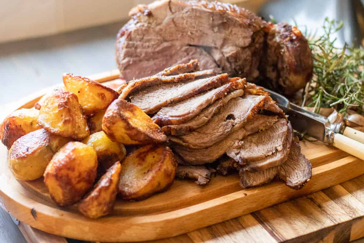 Sliced roast meat and golden roasted potatoes are served on a wooden cutting board with a knife and herbs in the background.