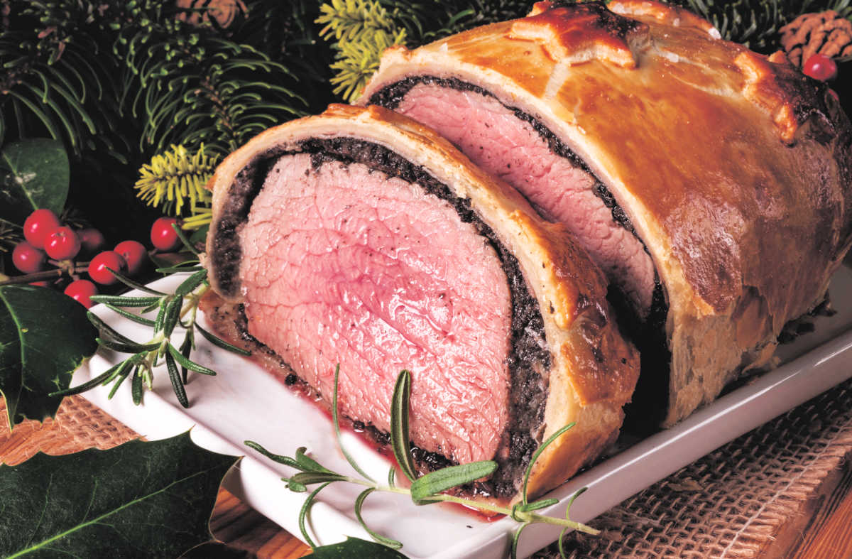 Sliced beef Wellington on a white platter, garnished with rosemary and surrounded by festive greenery and berries.