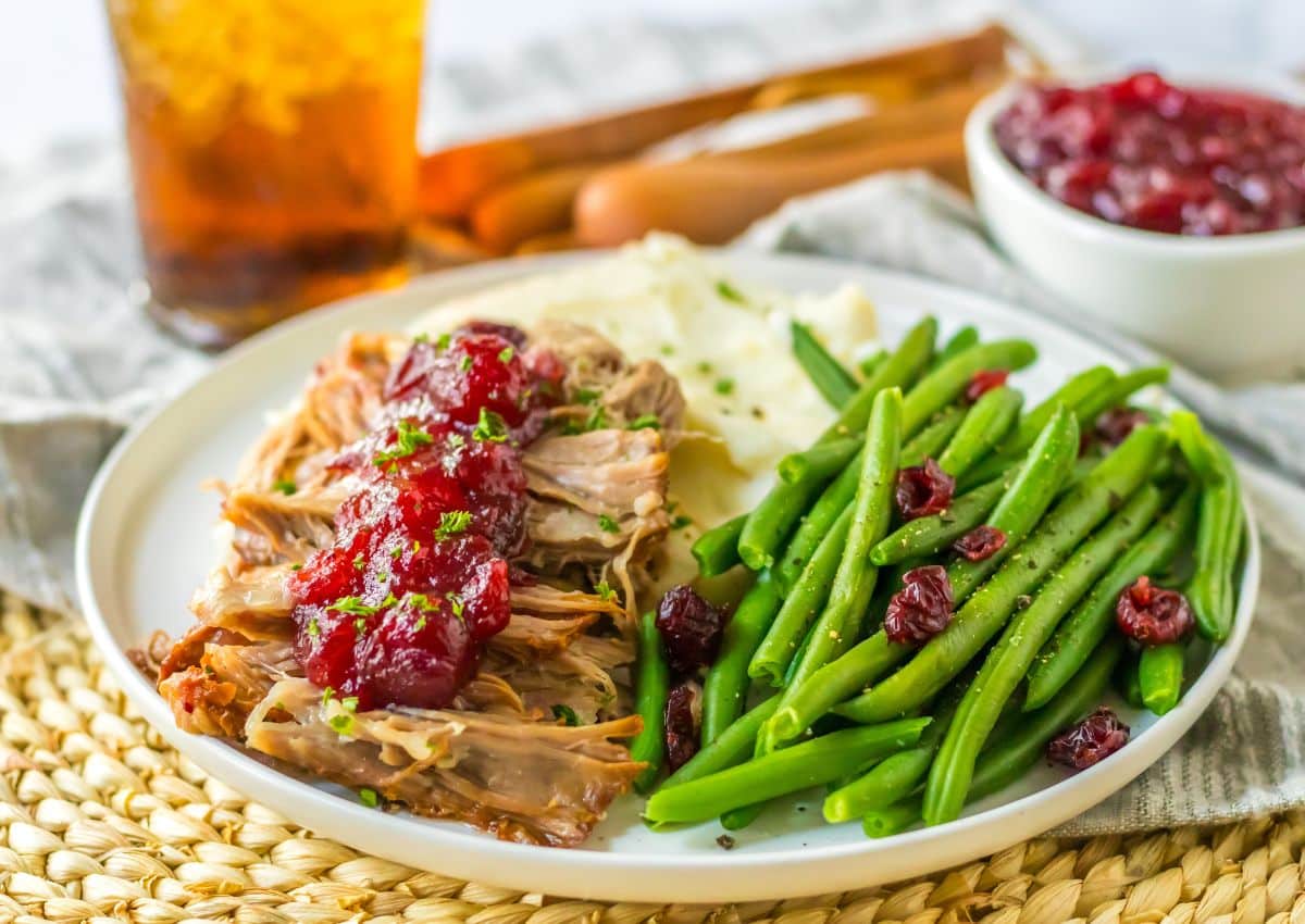 A plate with shredded roast meat topped with red sauce, mashed potatoes, green beans with cranberries, and a drink in the background.