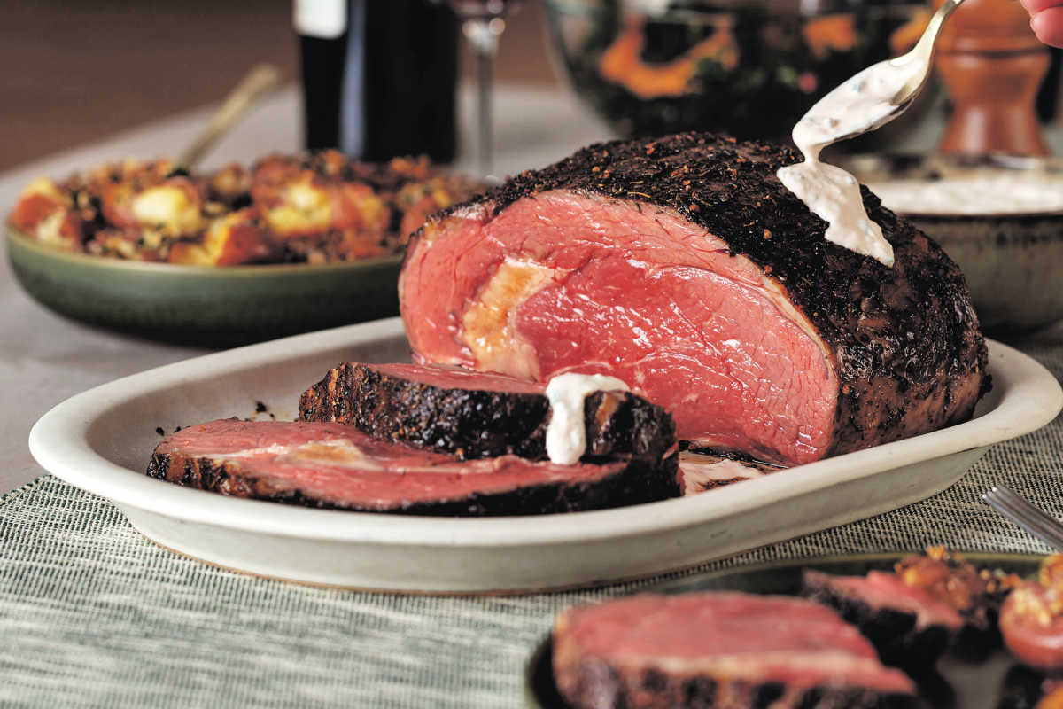 A sliced roast beef with a pink center is served on a platter, with creamy sauce being spooned over the meat. Sides are visible in the background.