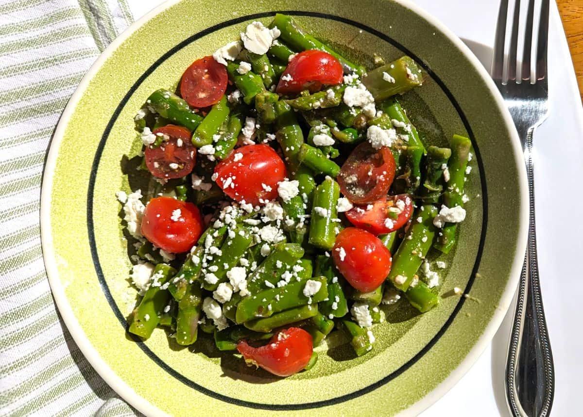 A bowl of green bean salad with halved cherry tomatoes and crumbled feta cheese, placed on a table with a striped napkin and a fork nearby.