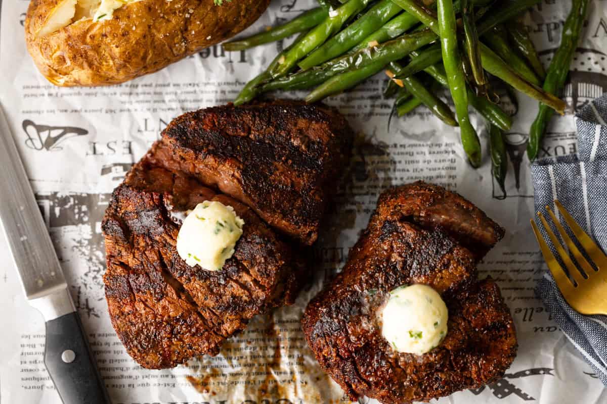 Two grilled steaks with herb butter on top, served with green beans and a baked potato, placed on newspaper. A knife, fork, and blue napkin are beside the food.