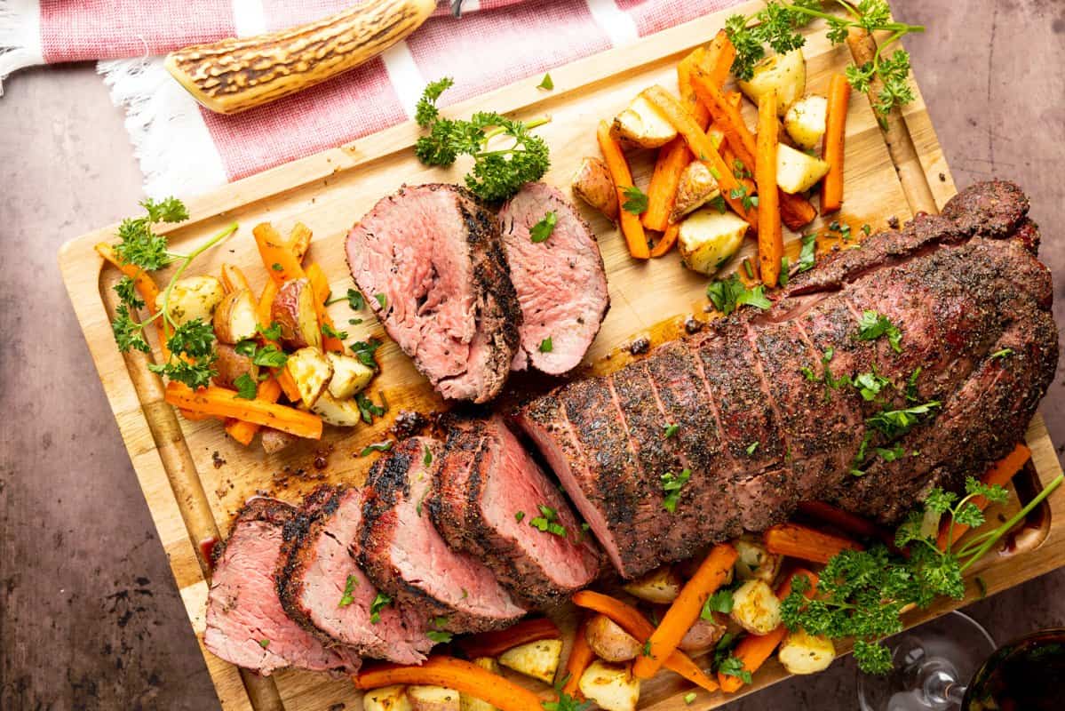 Sliced roast beef tenderloin garnished with herbs, served on a wooden board with roasted carrots and potatoes.
