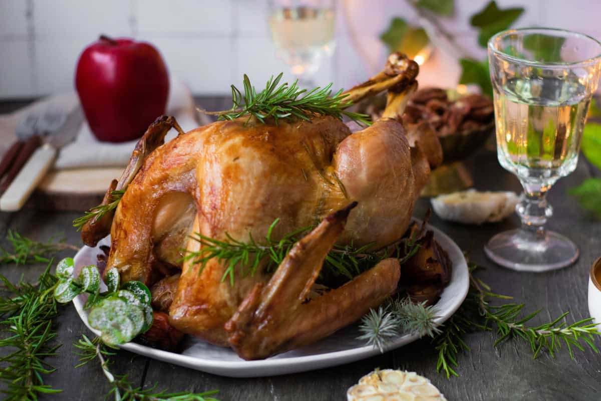 A roasted turkey garnished with fresh rosemary is served on a white plate, surrounded by glasses of white wine and an apple on a table.