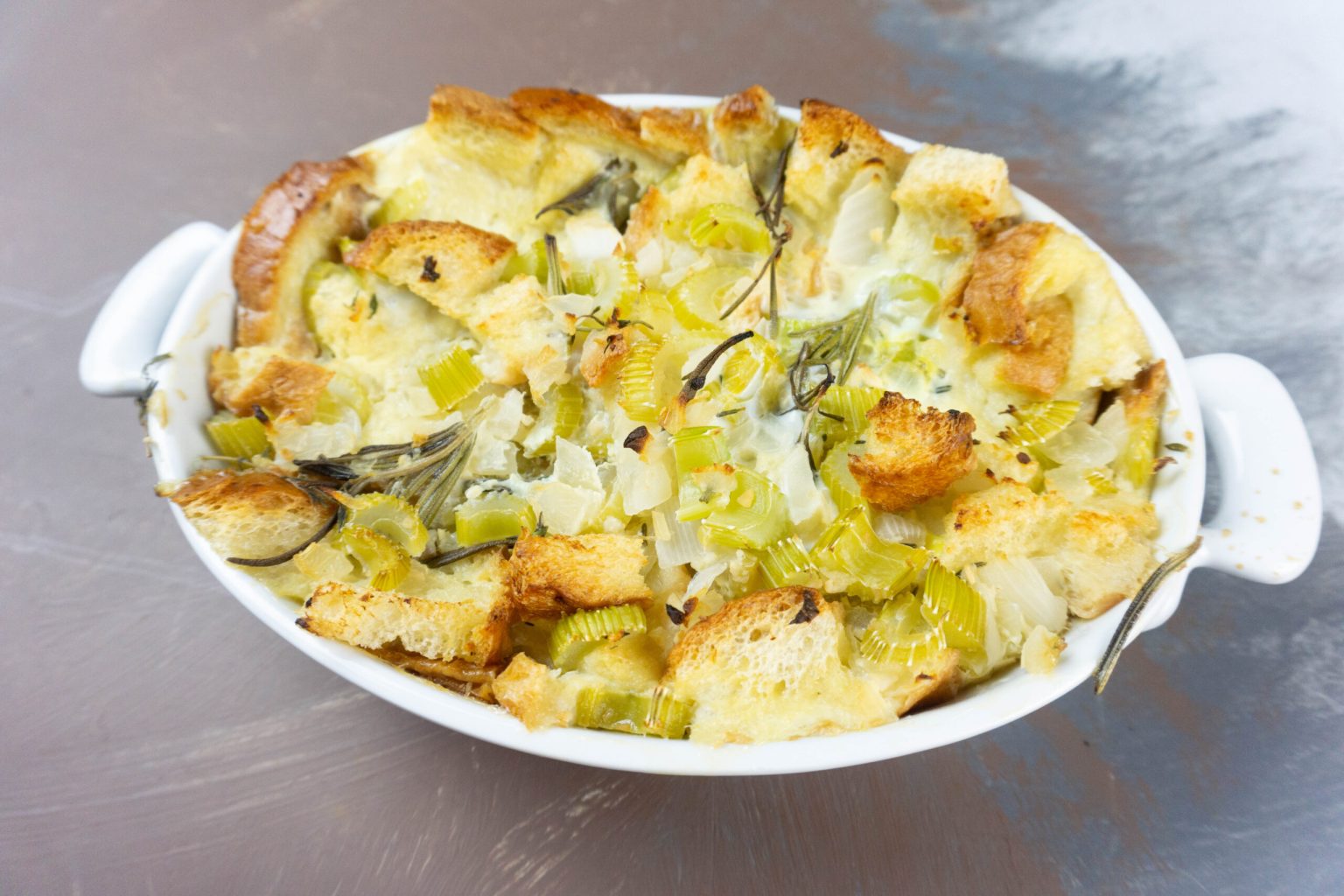 A white oval baking dish filled with baked bread cubes, celery, onions, and herbs, forming a savory casserole.