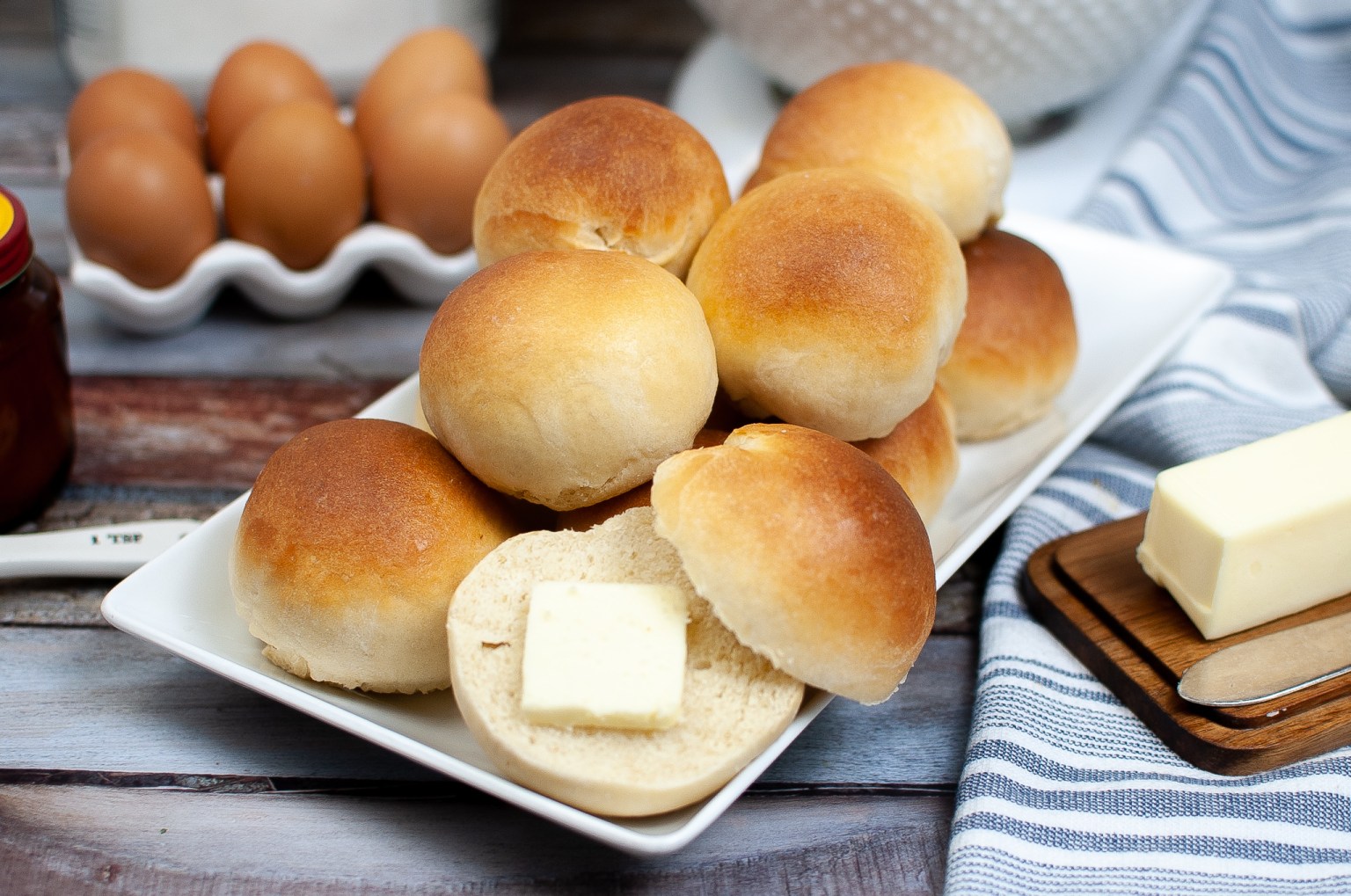 A plate of round bread rolls, with one roll cut open and topped with a slice of butter. Eggs, a butter stick, and a jar are in the background.