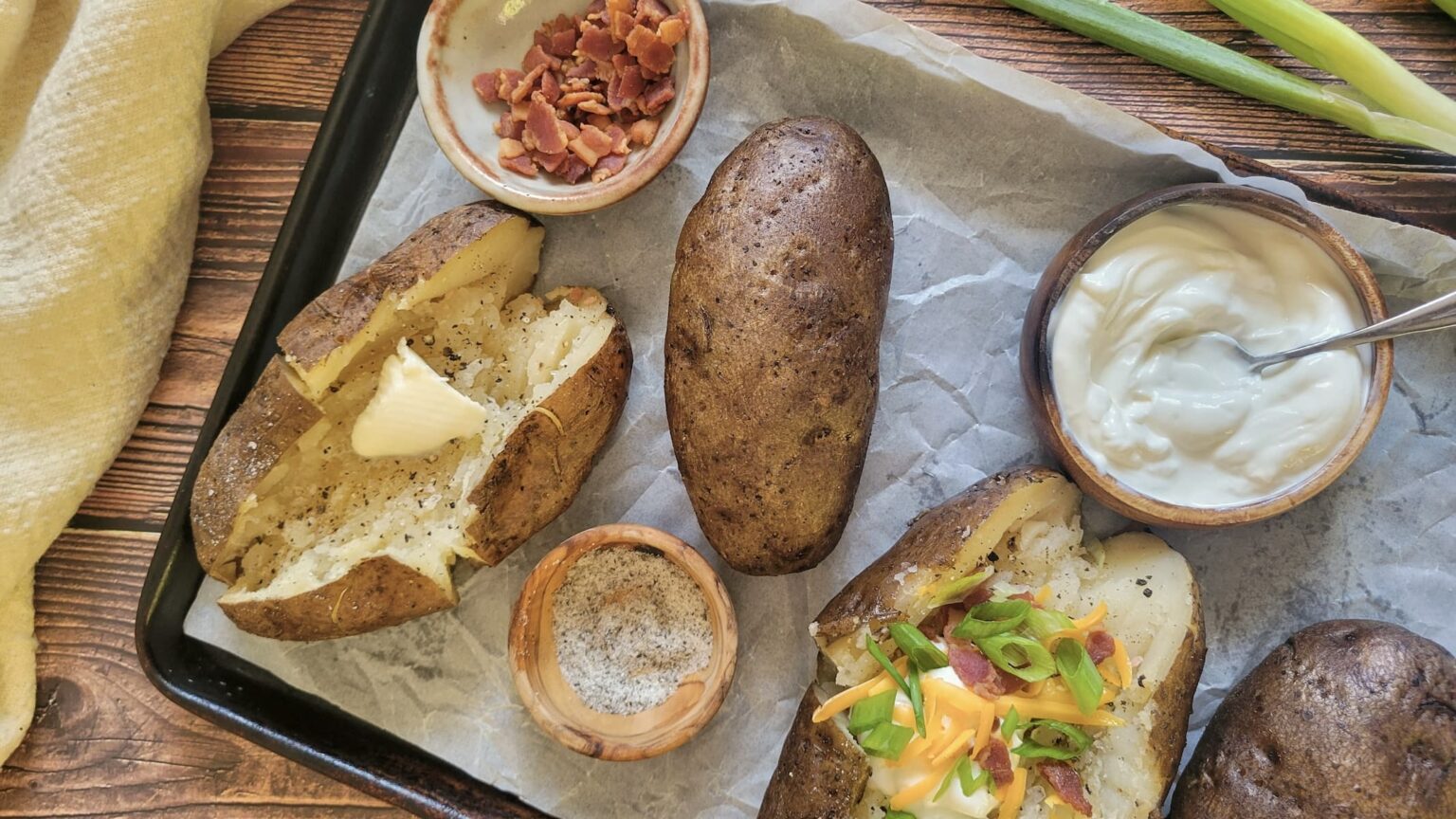 Baked potatoes on a tray with toppings including butter, sour cream, shredded cheese, bacon bits, sliced green onions, and a small bowl of salt.