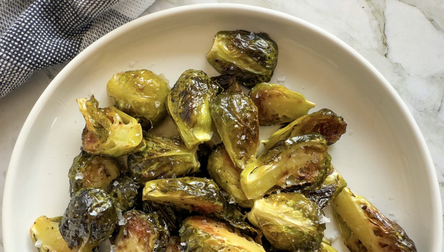 A white plate filled with roasted Brussels sprouts, lightly browned, sits on a marble surface next to a black and white cloth napkin.