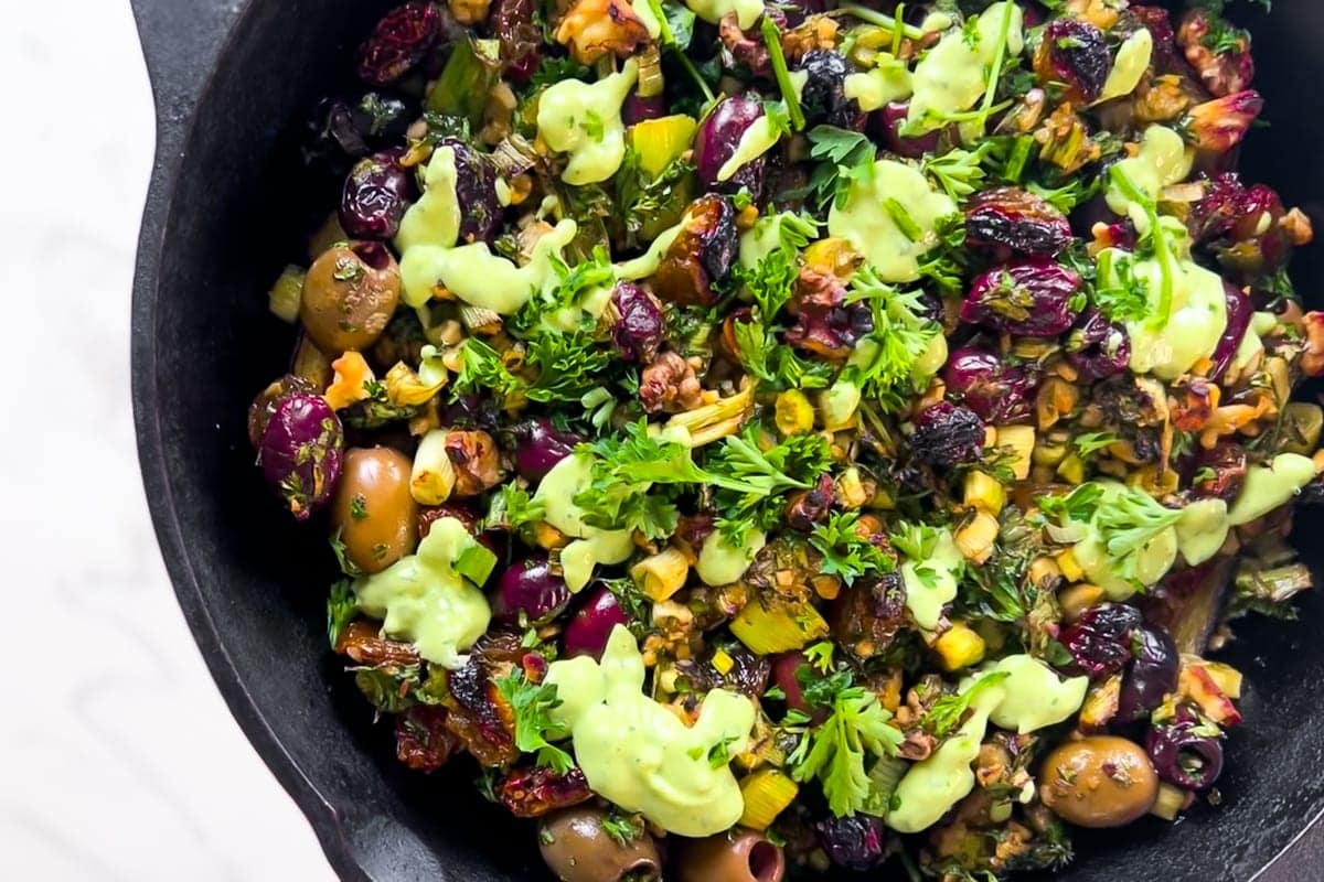 A cast iron skillet filled with a colorful mixture of olives, dried cranberries, herbs, nuts, and drizzled with a light green creamy sauce.