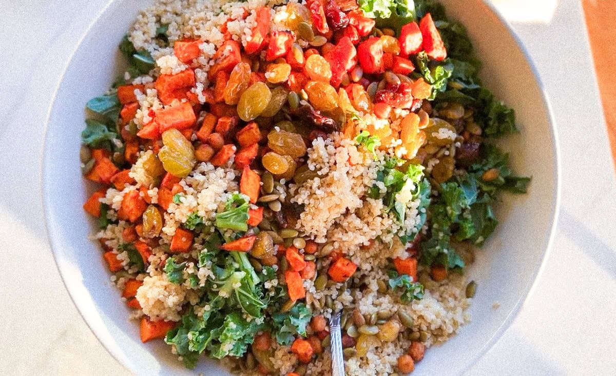 A bowl of quinoa salad with kale, roasted sweet potatoes, golden raisins, and pumpkin seeds, with a fork in the bowl.