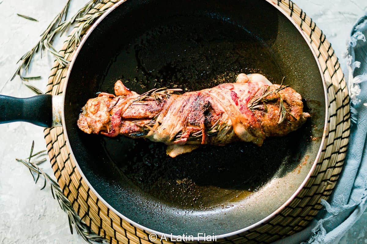 A cooked pork tenderloin wrapped in bacon and garnished with rosemary sits in a frying pan on a woven mat.