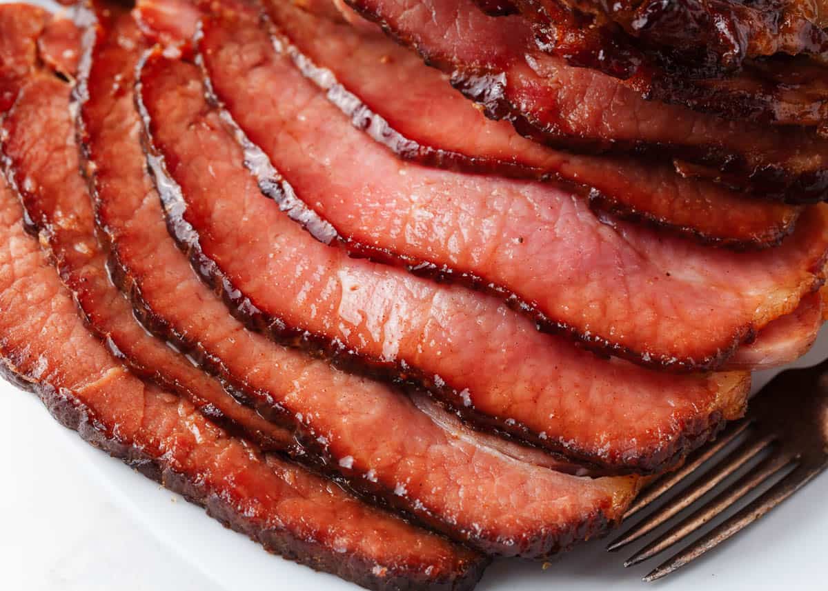 Close-up of several slices of glazed ham arranged on a plate with a fork visible in the bottom right corner.