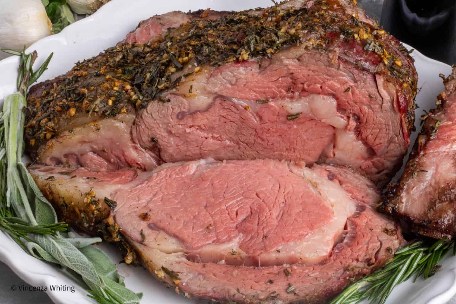 Close-up of a sliced, herb-crusted prime rib roast on a white platter, garnished with fresh rosemary and sage.