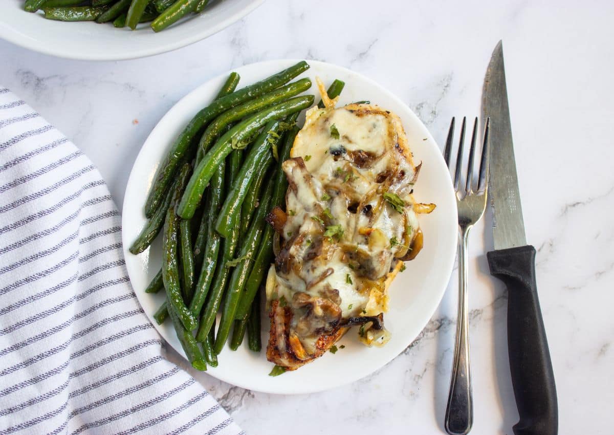 A plate with sautรฉed green beans and a piece of chicken topped with melted cheese and mushrooms, next to a fork and knife on a marble surface.