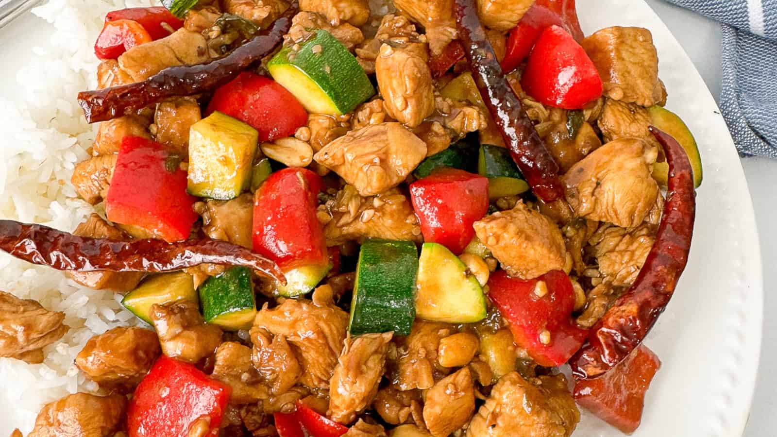 A plate of stir-fried chicken with zucchini, red bell pepper, dried red chilies, and white rice.