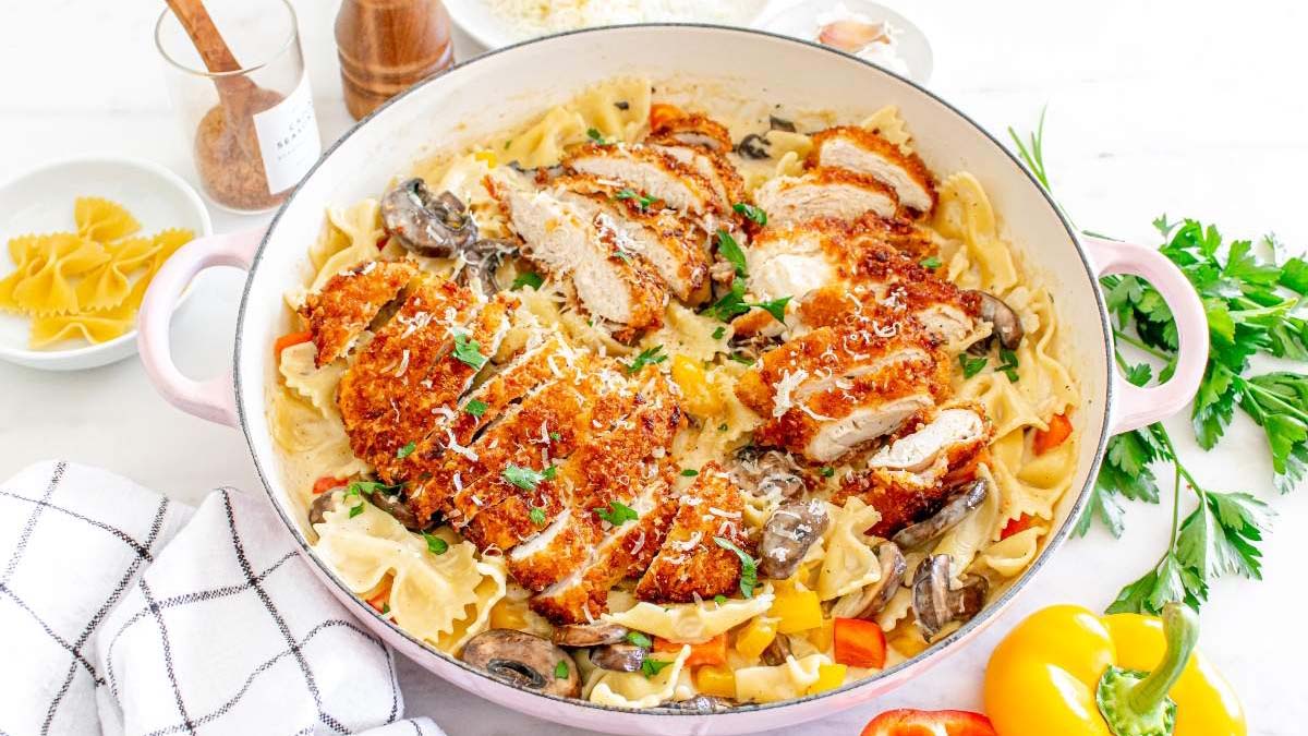 A pan of farfalle pasta with creamy sauce, sautรฉed mushrooms, bell peppers, and sliced breaded chicken cutlets, garnished with herbs and grated cheese.