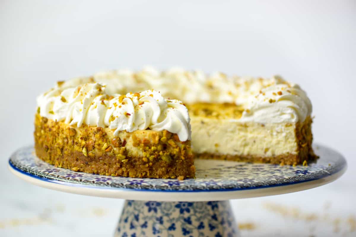 A cheesecake with a pistachio crust and whipped cream topping sits on a patterned cake stand, with one slice removed to show the creamy interior.