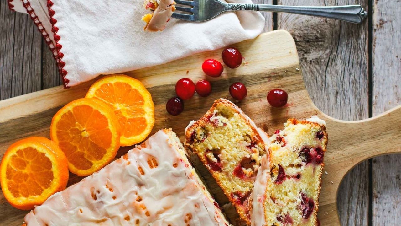 A loaf of glazed bread with cranberries, sliced on a wooden board, alongside orange slices and whole cranberries; a fork and napkin rest nearby.