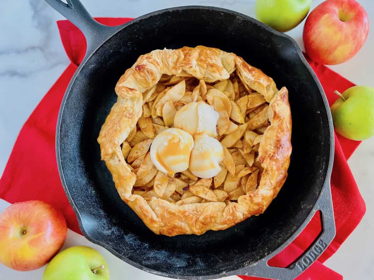 A baked apple galette topped with three scoops of vanilla ice cream in a black cast iron skillet, surrounded by fresh apples on a red cloth.