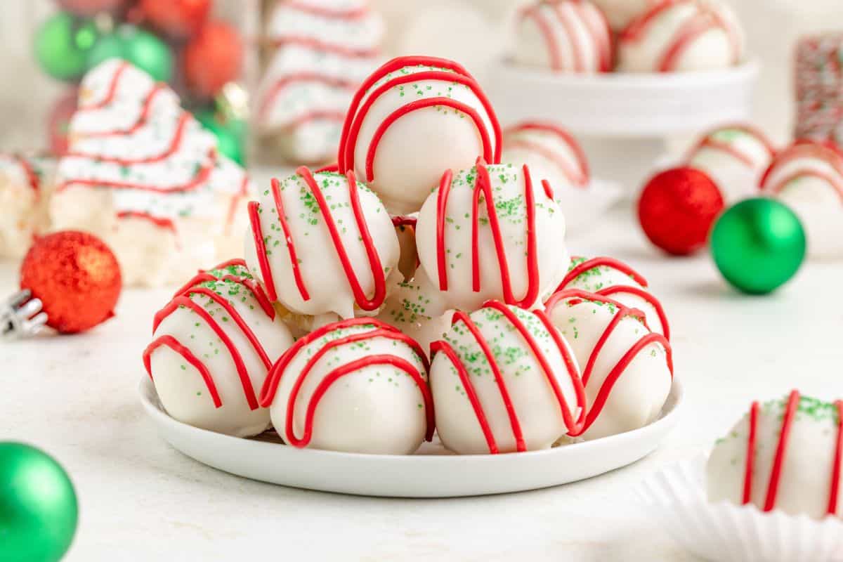 A plate of white chocolate-coated truffles decorated with red icing and green sprinkles, with Christmas ornaments and treats in the background.