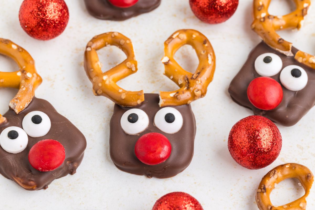 Chocolate-covered cookies decorated as reindeer with pretzel antlers, candy eyes, and red candy noses, surrounded by red glittery balls.