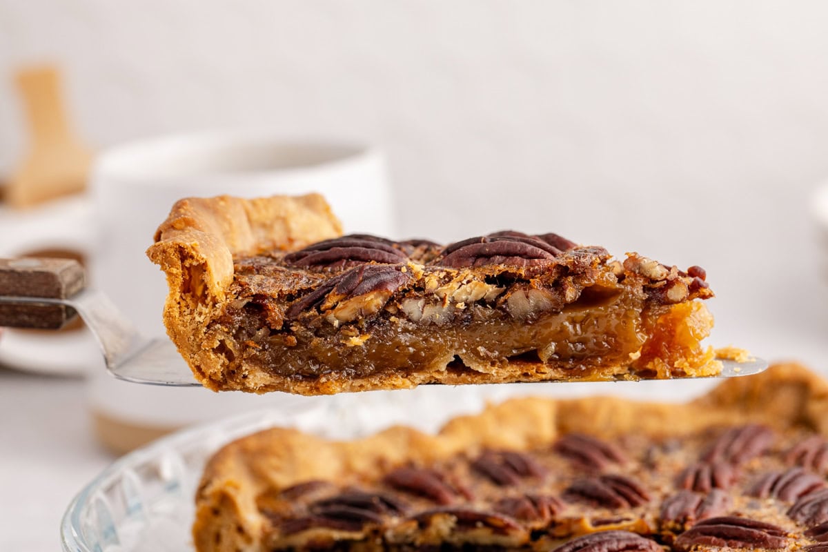 A slice of pecan pie being lifted from a pie dish, showing a flaky crust, gooey filling, and pecan topping.
