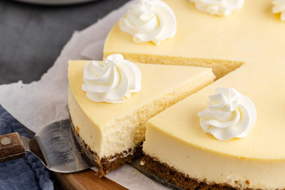 A cheesecake with a graham cracker crust is sliced, topped with swirls of whipped cream, and served on a wooden board with parchment paper.