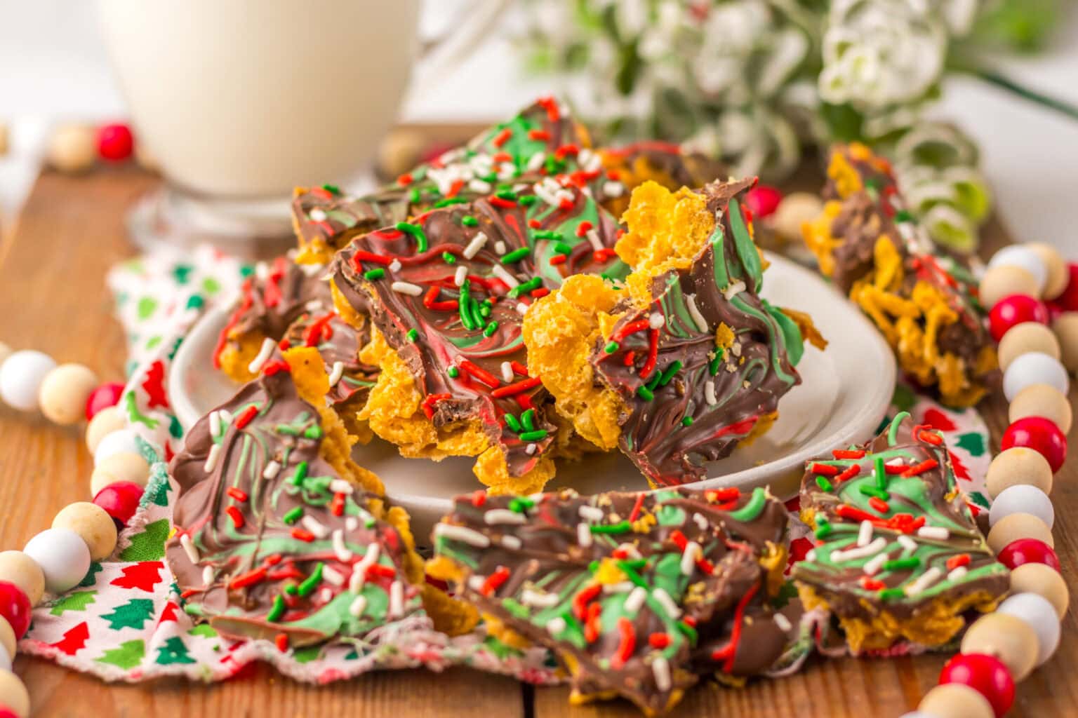 A plate of chocolate bark topped with red, green, and white sprinkles, set on a festive napkin with a glass of milk and holiday beads in the background.