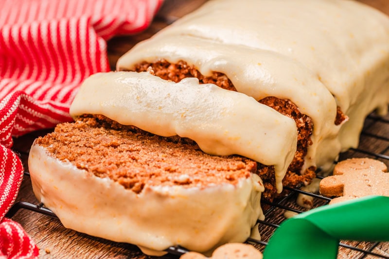 A loaf of gingerbread cake with creamy frosting, partially sliced, sits on a cooling rack next to a red striped cloth.