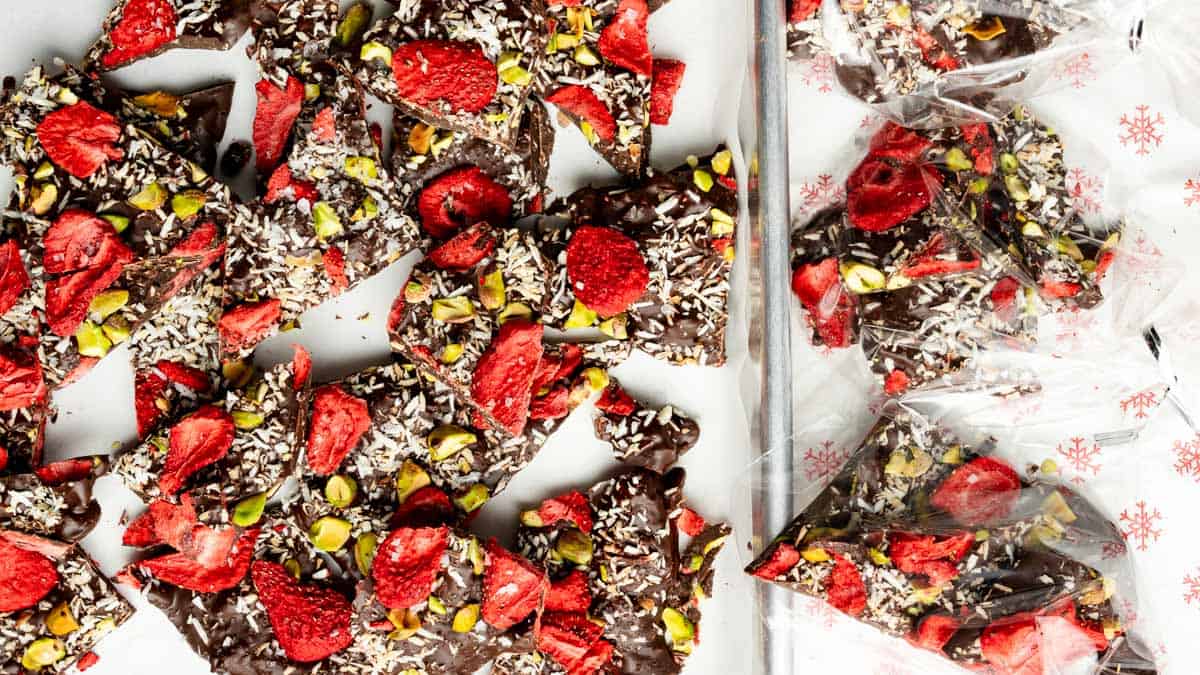 Broken pieces of dark chocolate bark topped with dried strawberries, pistachios, and shredded coconut, some pieces placed in a clear plastic bag with a snowflake pattern.