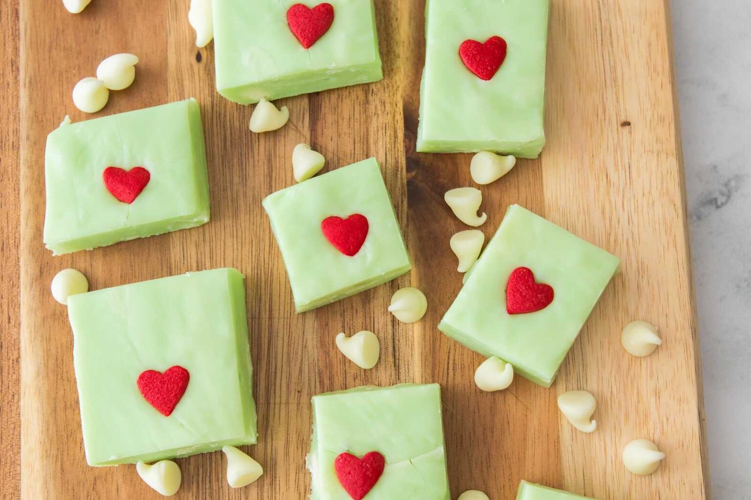 Squares of green fudge topped with red heart decorations are arranged on a wooden board with scattered white chocolate chips.