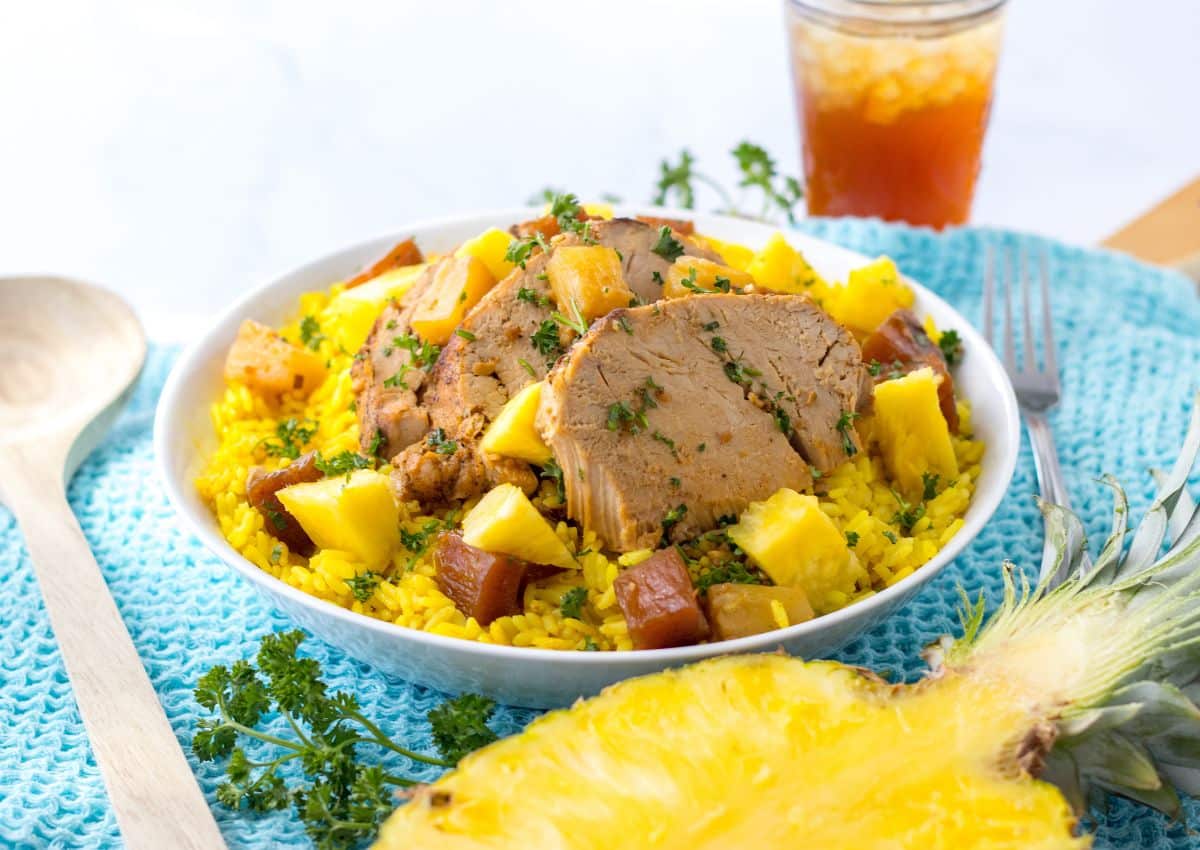 A bowl of yellow rice topped with sliced pork, pineapple chunks, and herbs sits beside a wooden spoon, a glass of iced tea, and a half pineapple.