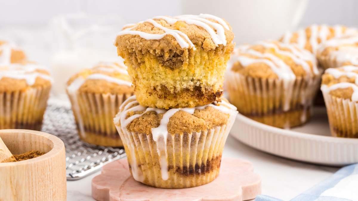 Two crumb-topped muffins with white icing drizzle are stacked on a pink coaster, with more muffins and baking items in the background.