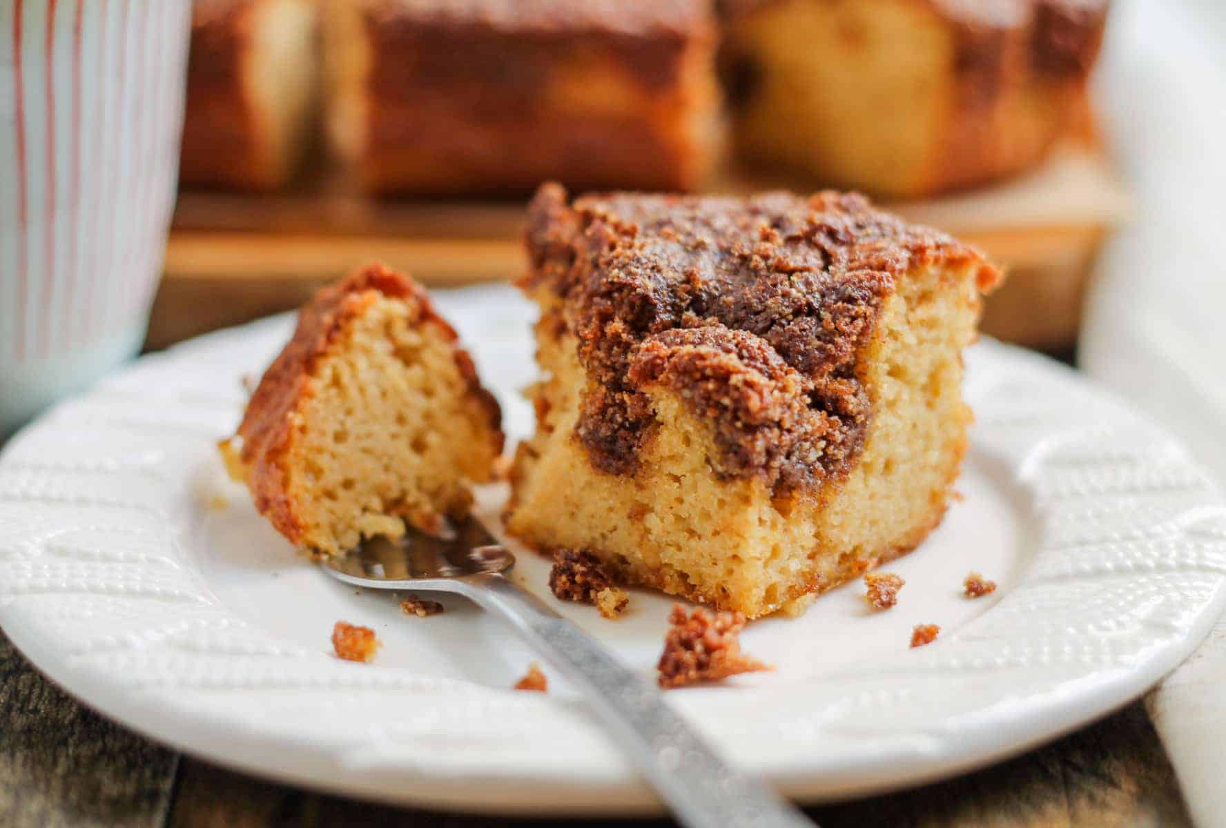 A slice of cinnamon coffee cake with a crumb topping sits on a white plate; a fork rests beside it with a piece of cake on the fork.