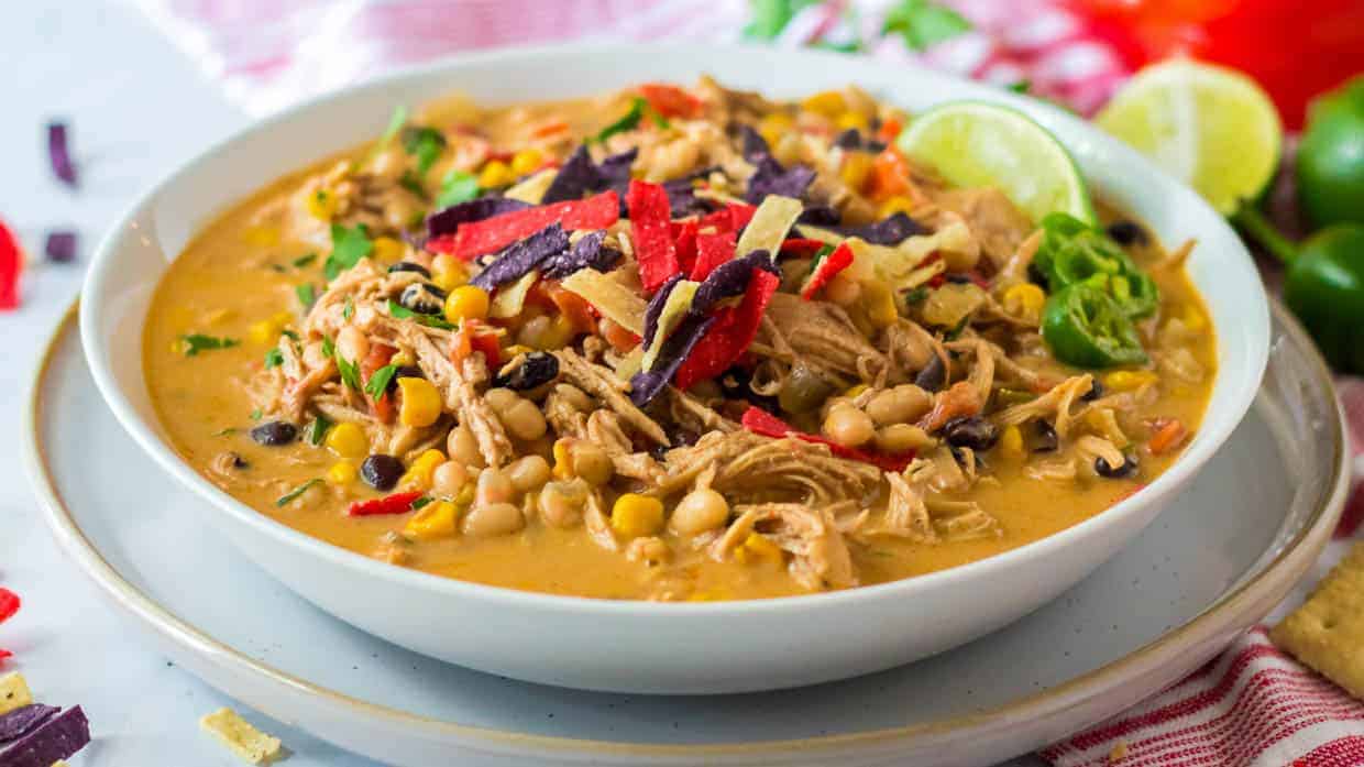 A bowl of creamy chicken chili topped with shredded tortilla strips, lime slices, and jalapeรฑos, containing corn, beans, and herbs.