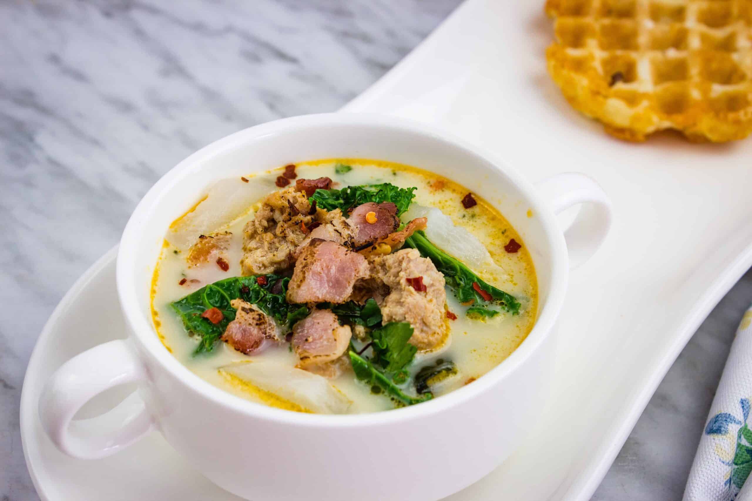 A white bowl of creamy soup with sausage, bacon, kale, and potatoes, served on a white tray next to a waffle.