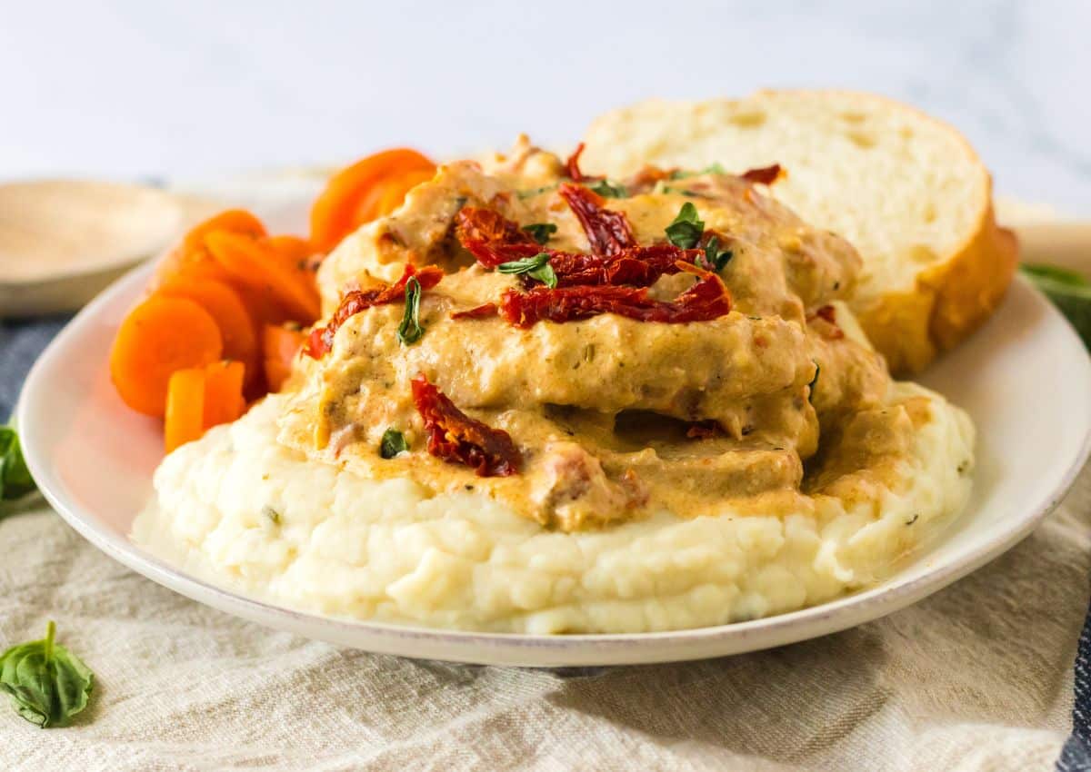A plate of mashed potatoes topped with creamy chicken and sun-dried tomatoes, served with cooked carrots and a slice of bread.