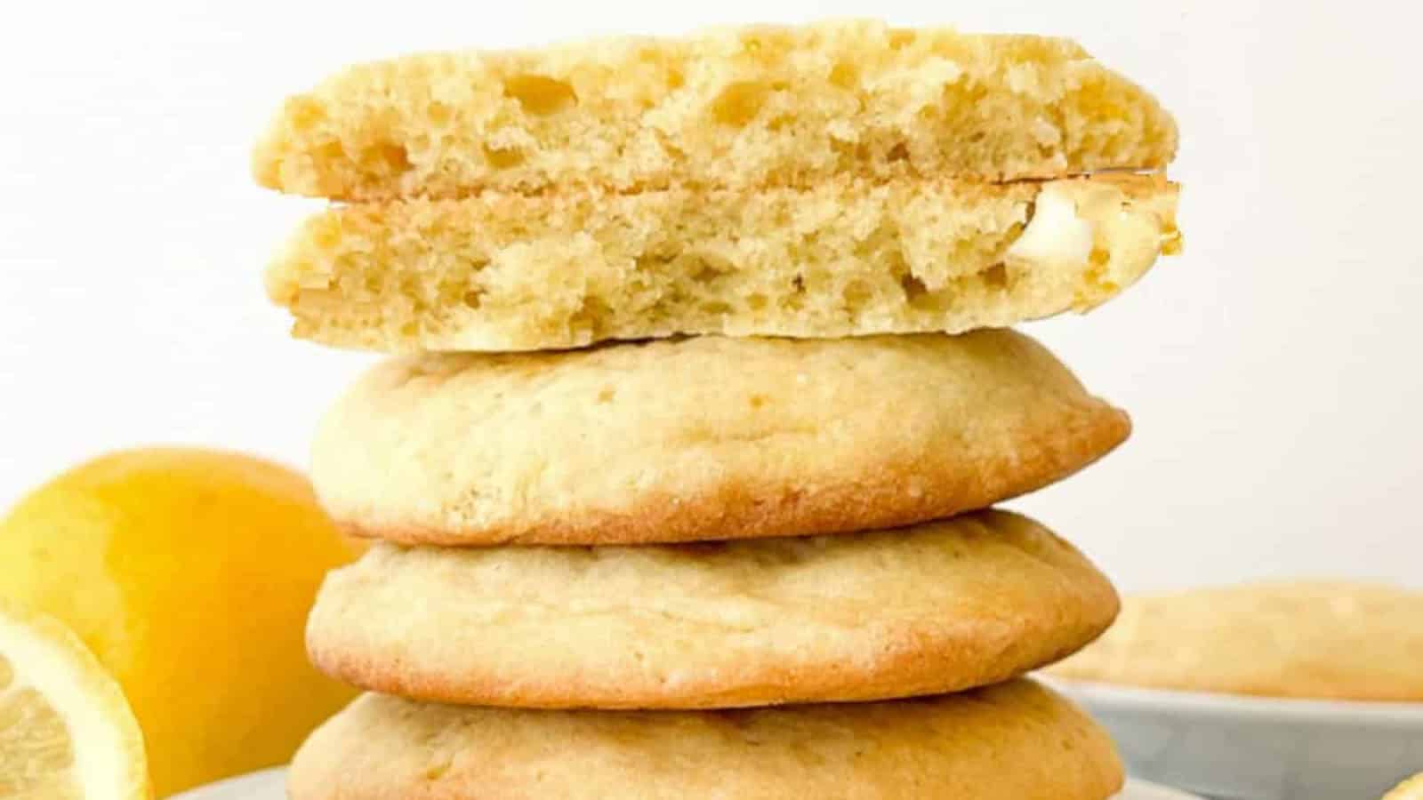 A stack of three lemon cookies with a partially broken cookie on top, showing the soft, fluffy interior. A lemon is visible in the background.