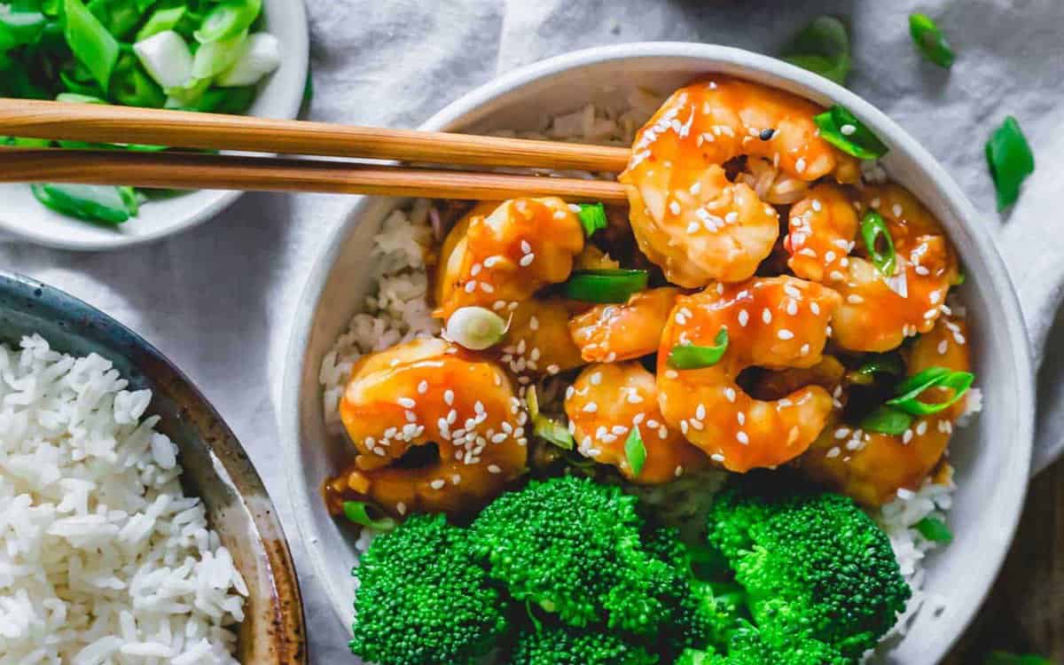 A bowl of rice topped with glazed shrimp, sesame seeds, and chopped green onions, served with steamed broccoli; extra rice and green onions are on the side.