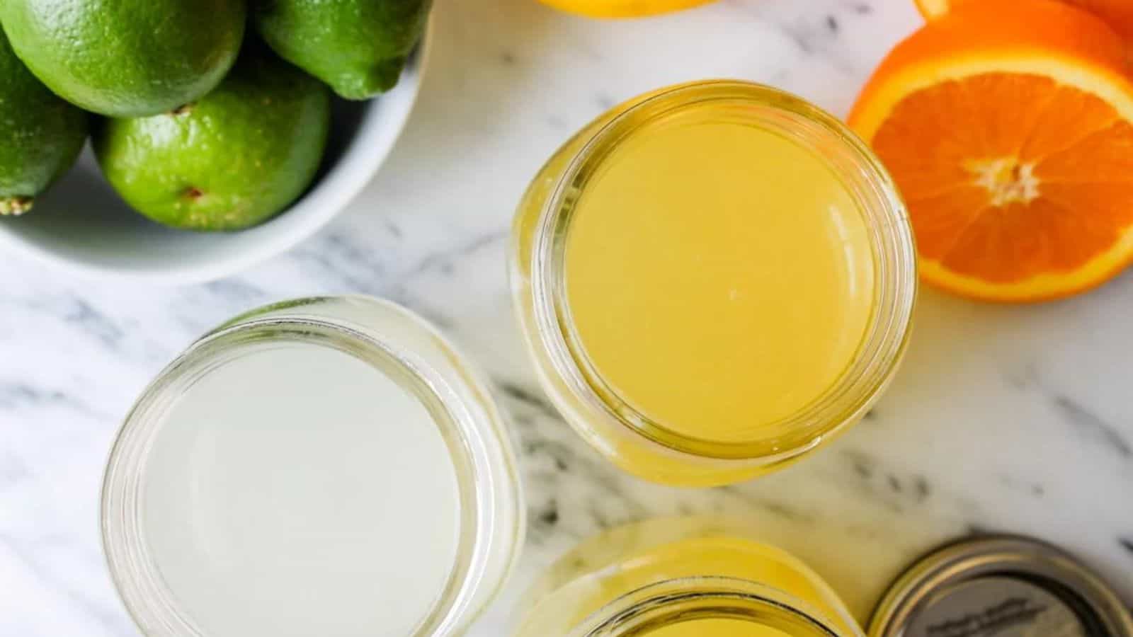 Three jars of citrus juice on a marble surface, with whole limes in a bowl and halved oranges nearby.